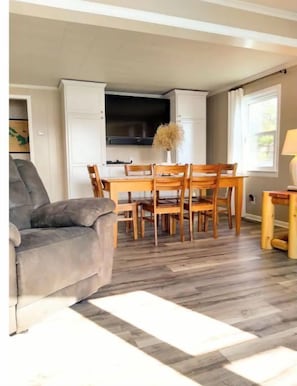 A living room with a grey sofa, a dining area with wooden chairs and a table, and a kitchen with white cabinets and a TV.