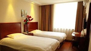 Une chambre d’hôtel avec deux lits simples, une tête de lit en bois, une table de chevet avec une lampe, un vase de fleurs et une vue sur l’extérieur par une fenêtre avec des rideaux.