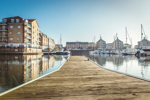 Une jetée en bois menant à une marina où se trouvent des bateaux, des bâtiments et un ciel dégagé.