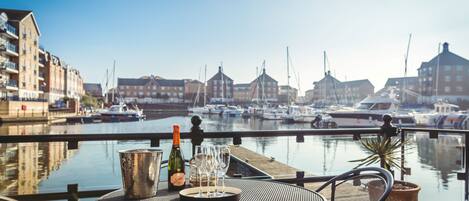 A table with a bottle and glasses, overlooking a marina with boats and buildings.
