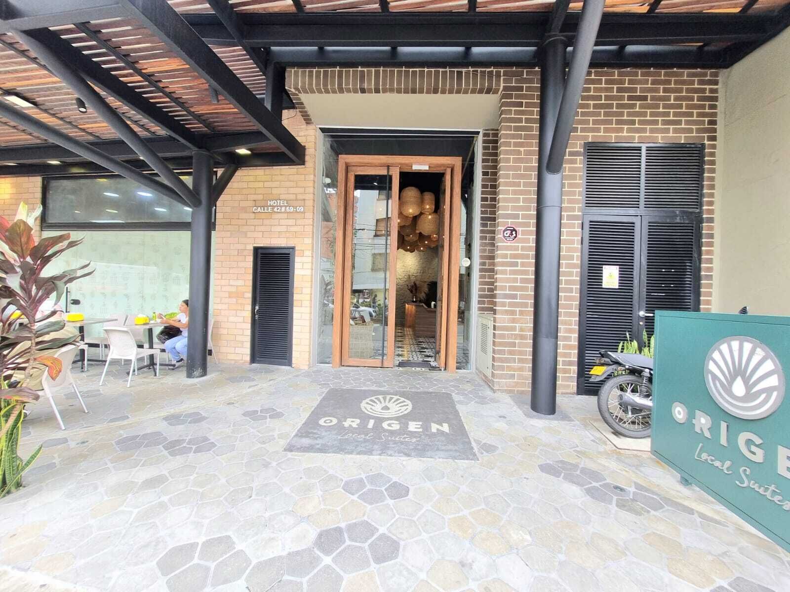 Een hotelentree met een betegeld terras, een bordje met 'ORIGEN Local Suites' en een gevel van baksteen.