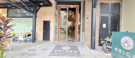 A hotel entrance with a tiled patio, a sign for 'ORIGEN Local Suites', and a brick building facade.
