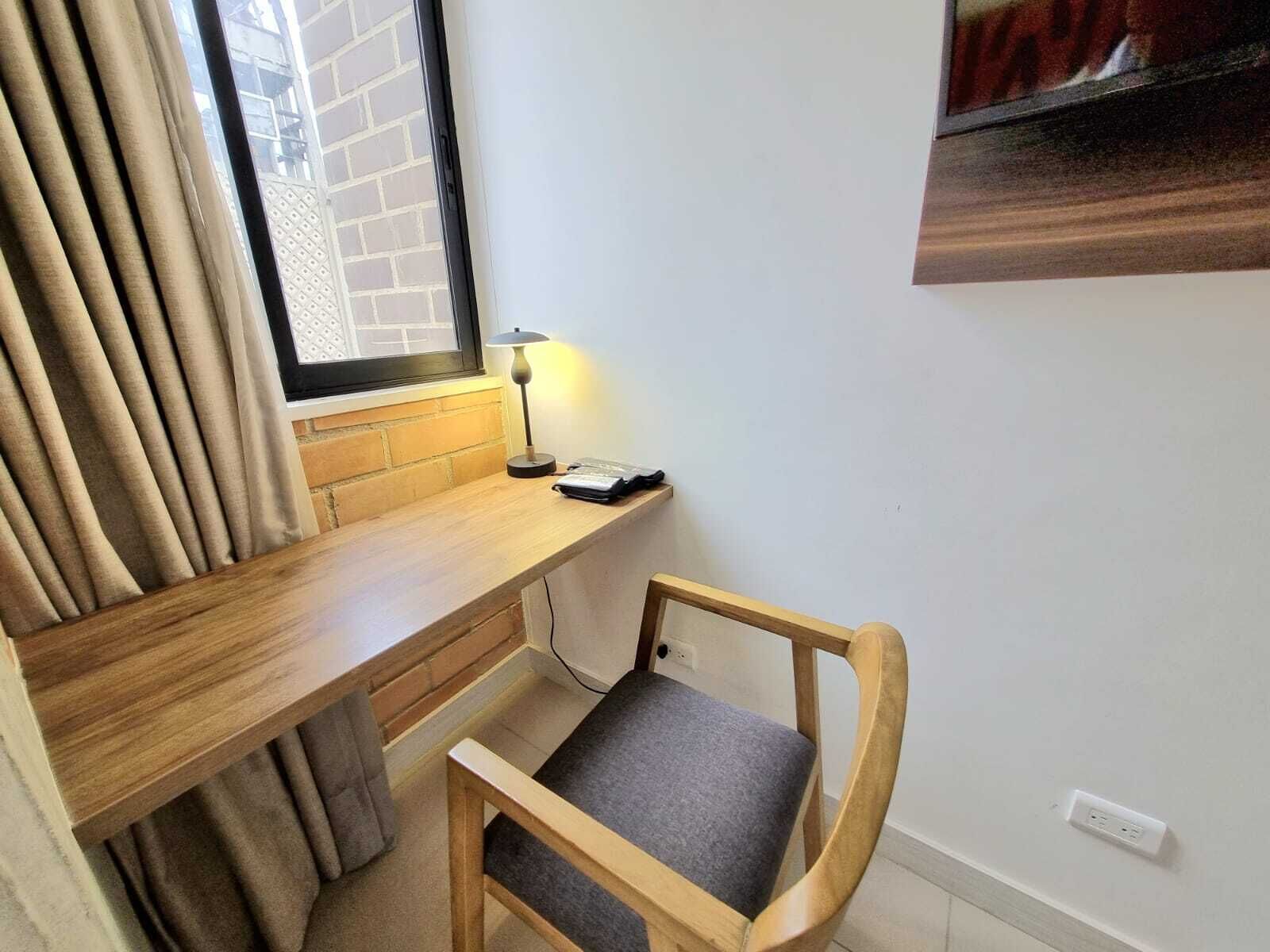 Un espacio de trabajo pequeño y ordenado, con un escritorio de madera, una silla, una lámpara y una ventana con persianas.