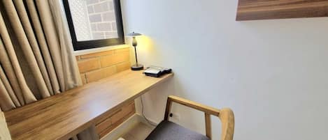 Un espacio de trabajo pequeño y ordenado, con un escritorio de madera, una silla, una lámpara y una ventana con persianas.