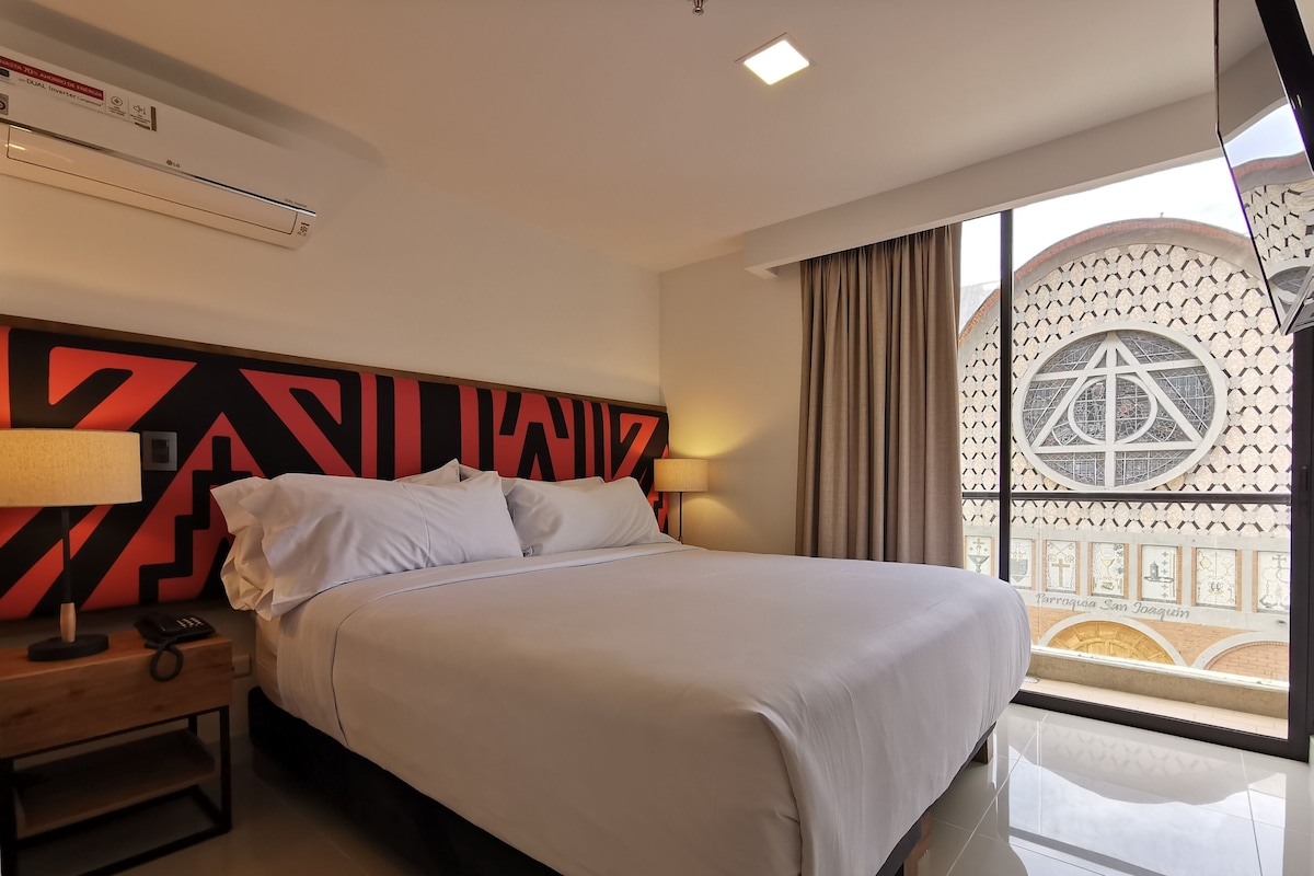A hotel room with a bed, bedside table, lamp, and a large window with a view of a building.
