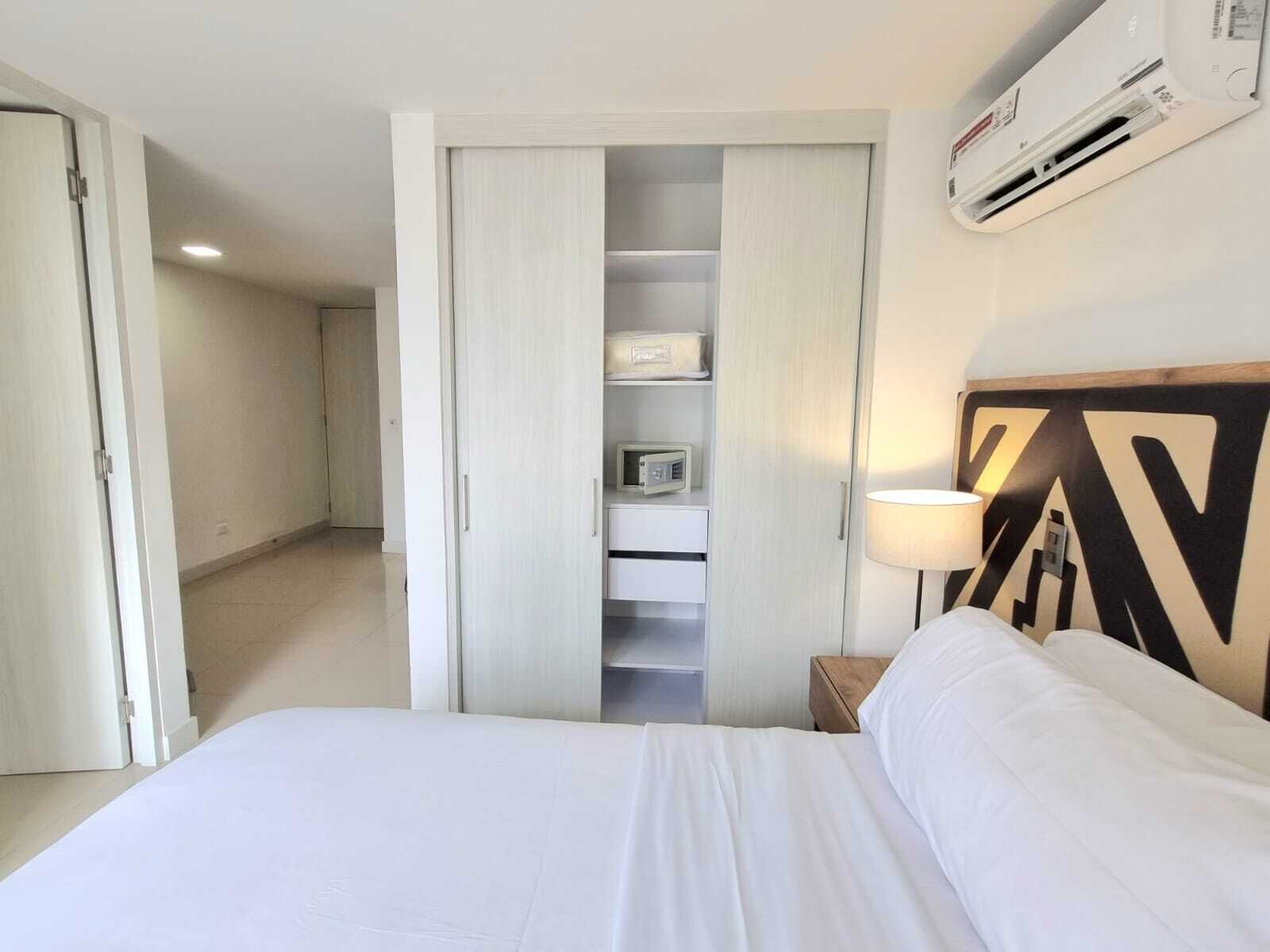 A hotel room with a bed, a wardrobe with a mirror, a bedside table with a lamp, and an air conditioning unit on the wall.