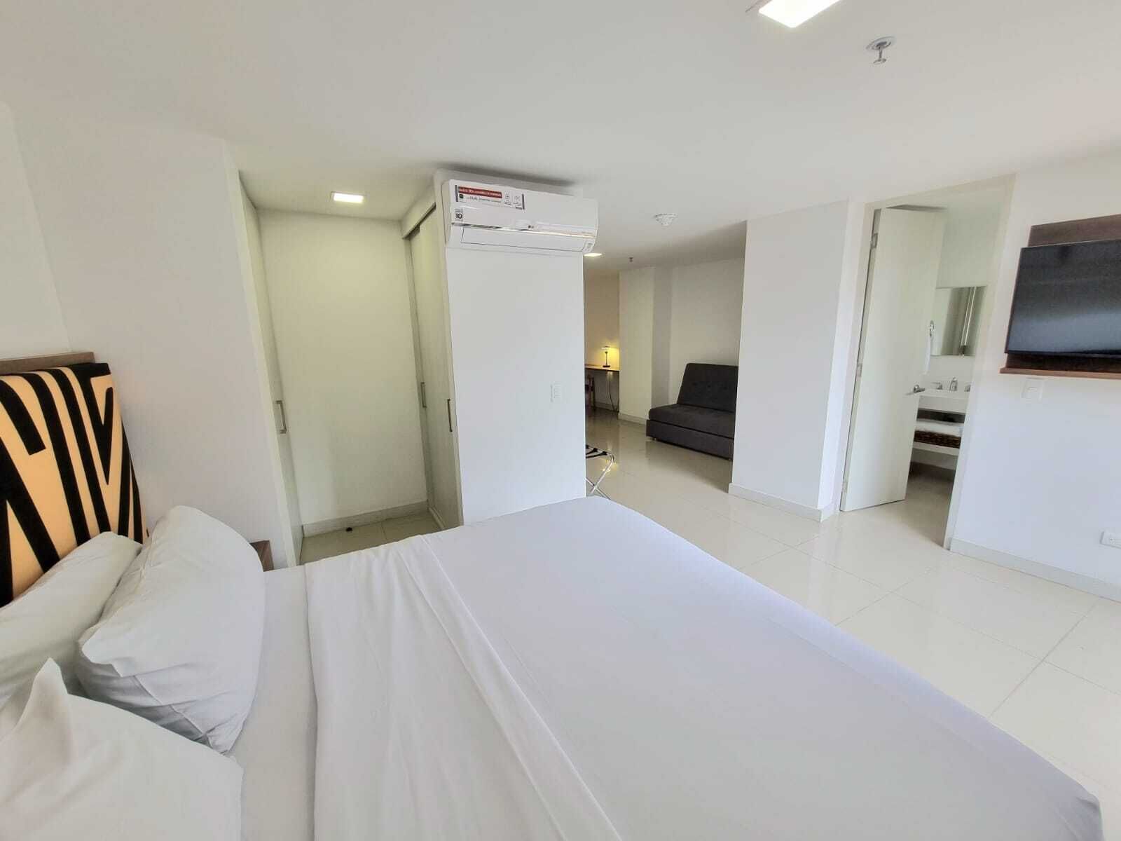 A hotel room with a bed, a sofa, a TV, and a bathroom visible through an open door.