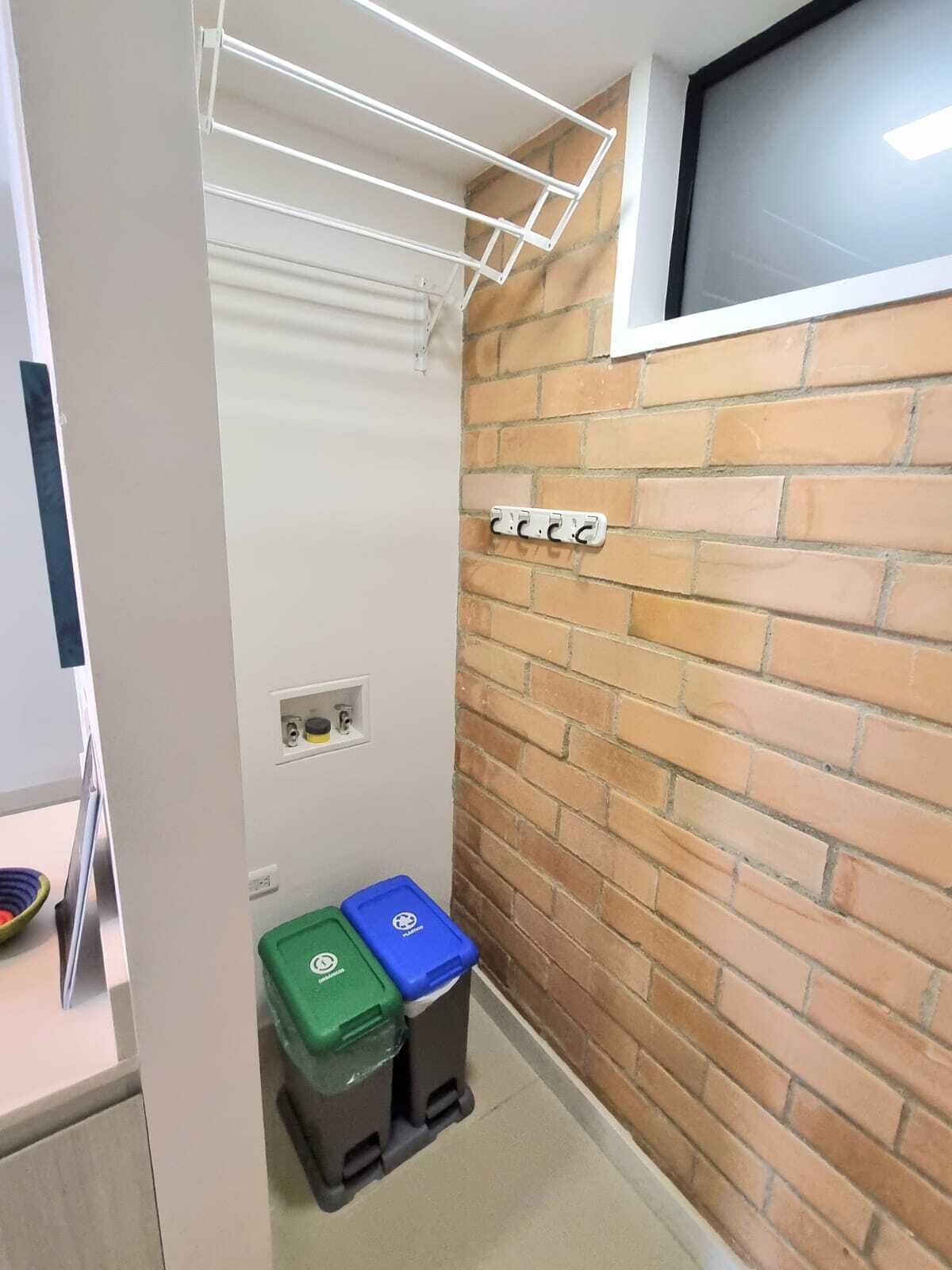 A small room with a brick wall, a window, a white shelf with a hanging rack, and two trash bins.