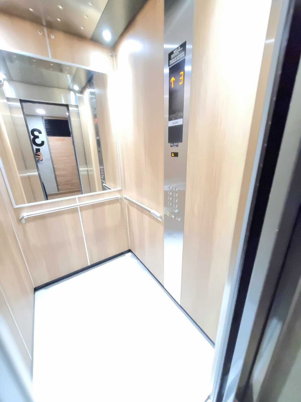 An elevator interior with a mirrored wall, a handrail, and a control panel with directional arrows.