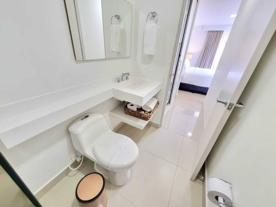 A modern bathroom with a toilet, sink, and mirror. There is a towel rack above the sink and a basket of toiletries on the countertop.