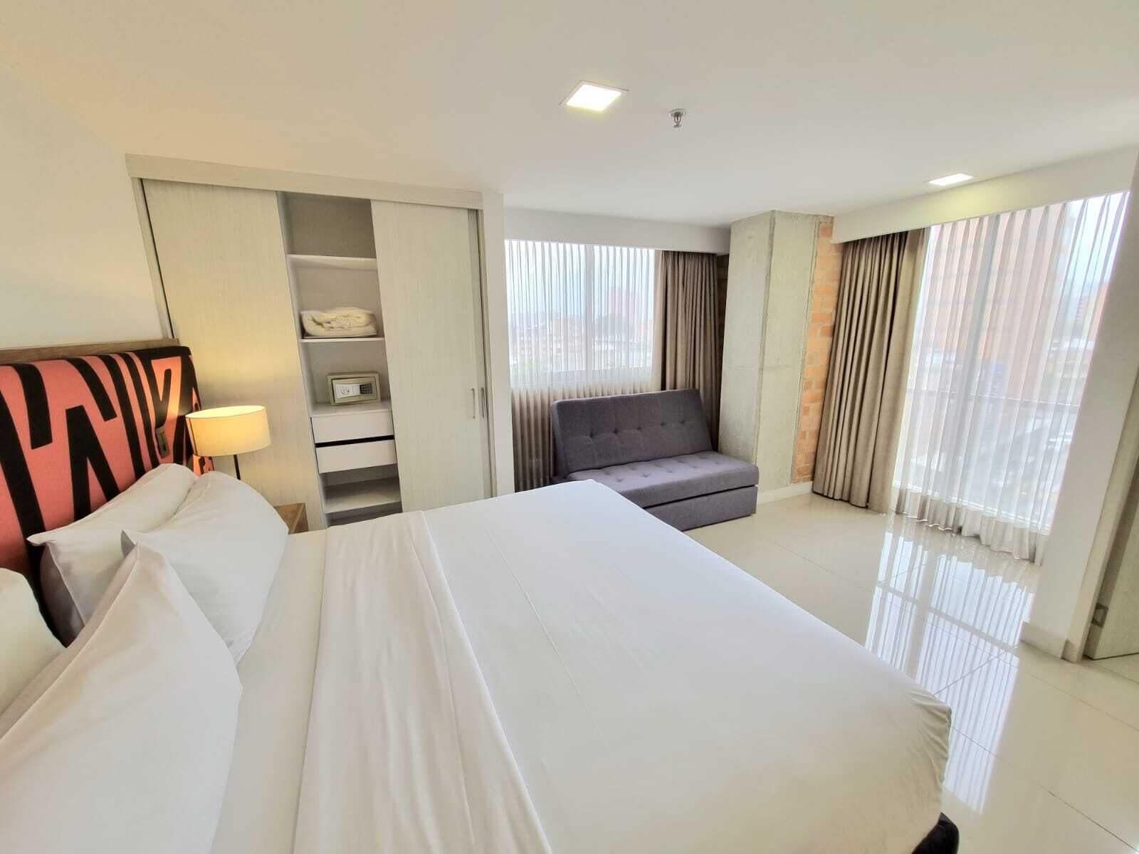 A hotel room with a large bed, a grey sofa, a wardrobe, and a window with curtains.