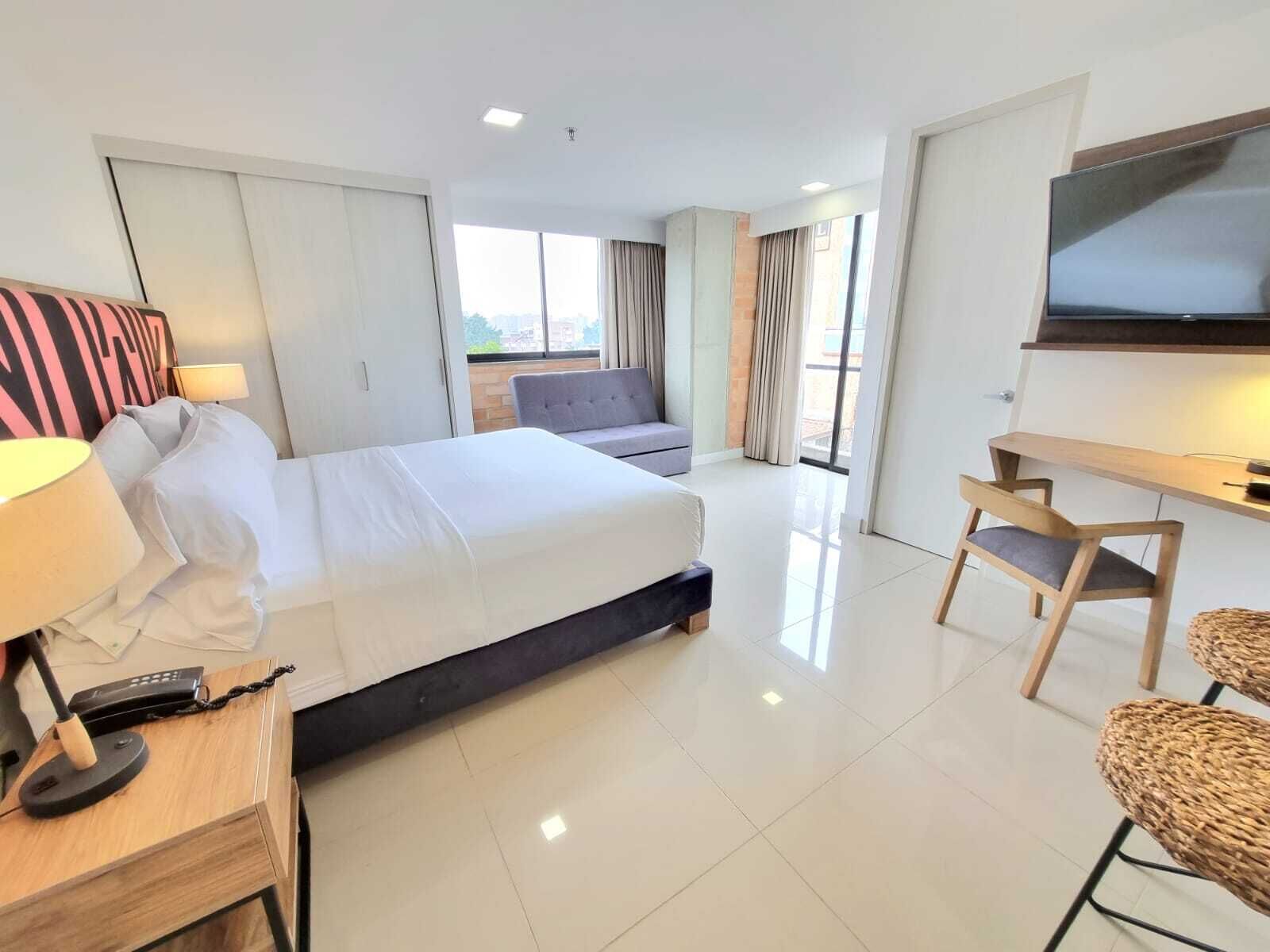 A hotel room with a bed, a desk with a lamp, a TV, and a seating area with a sofa and chairs.