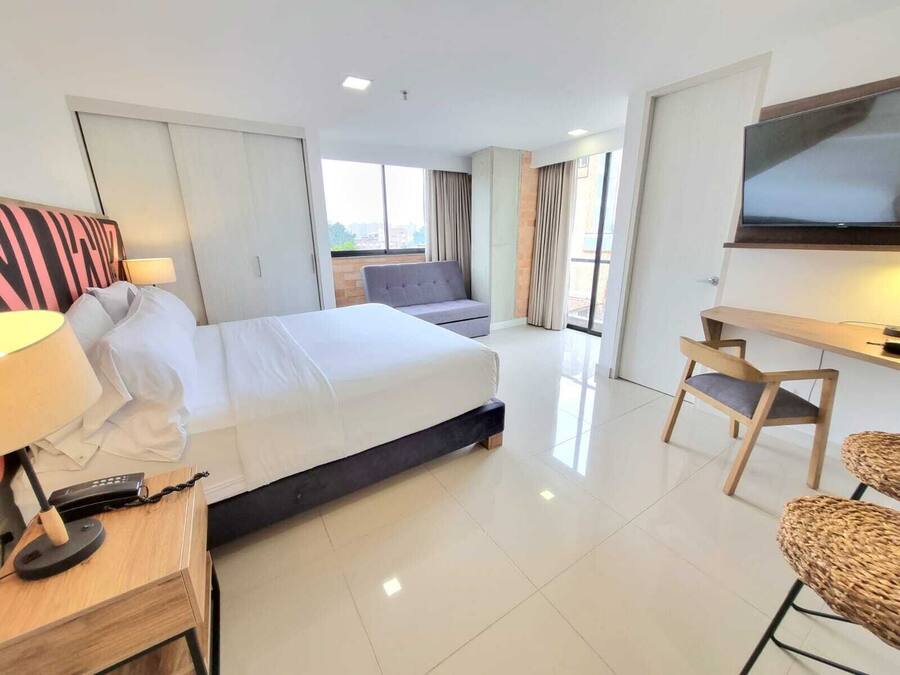 A hotel room with a bed, a desk with a lamp, a TV, and a seating area with a sofa and chairs.