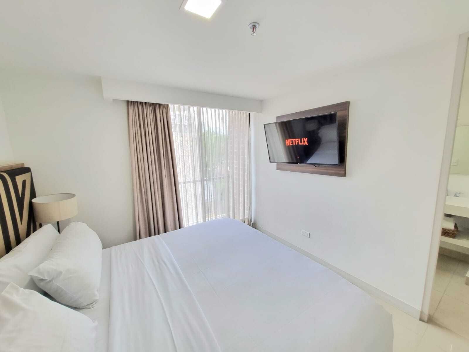 A hotel room with a bed, a TV mounted on the wall, a window with curtains, and a bedside table with a lamp.