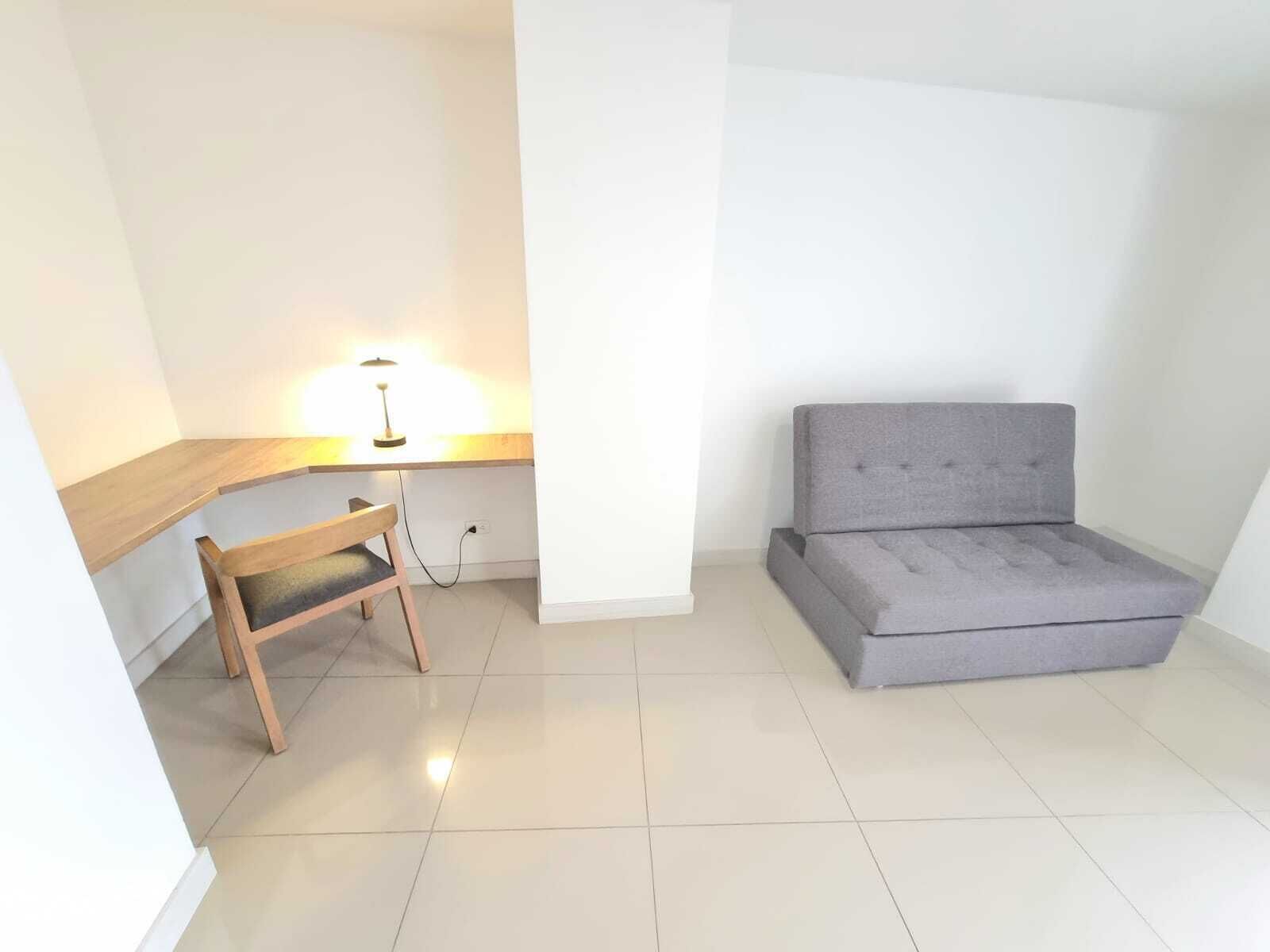 Ein kleiner, heller Raum mit einem Holztisch, einem grauen Sofa und einer Lampe.