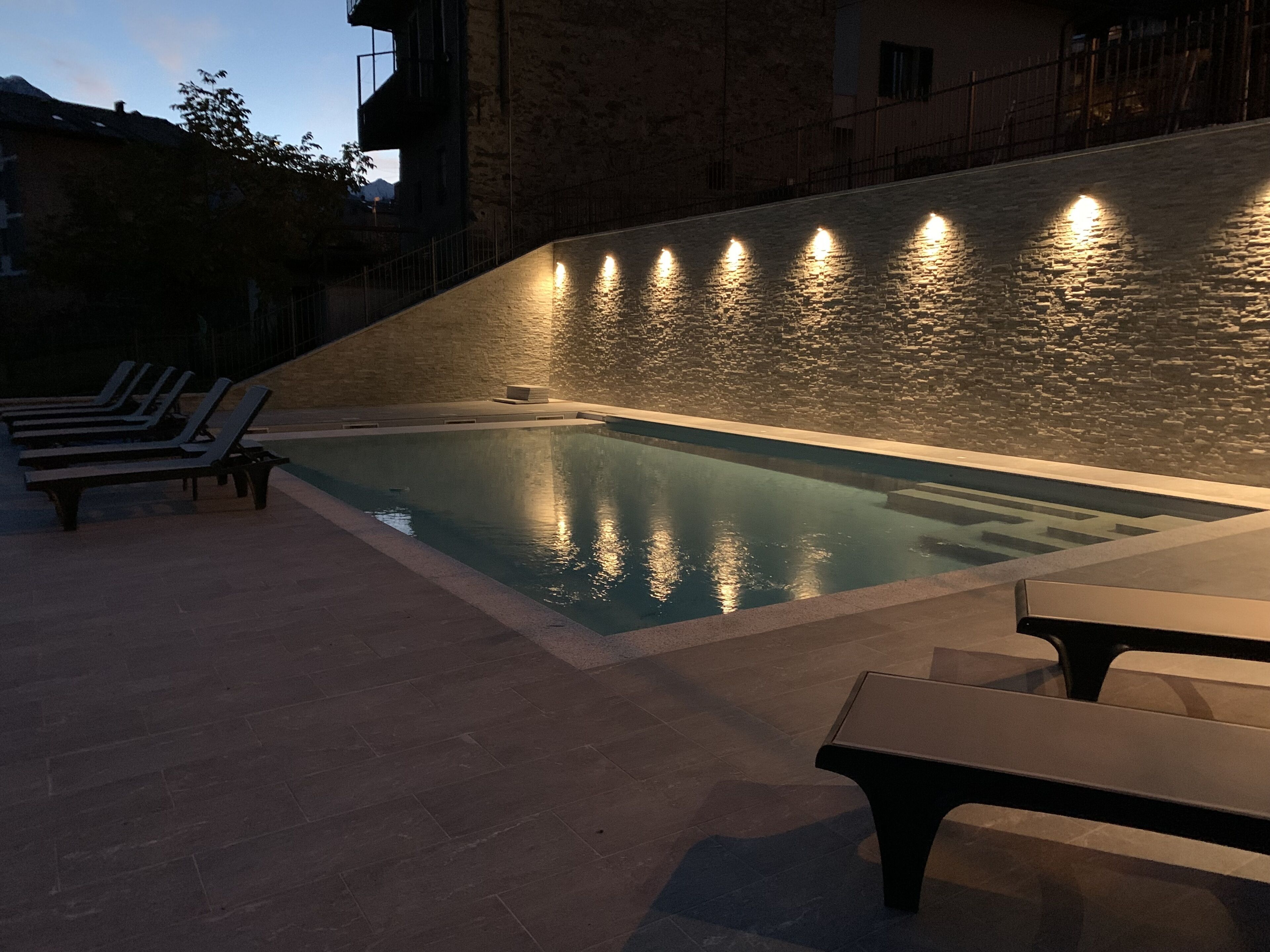 Une piscine avec un mur en pierre et des éclairages, entourée de chaises longues et d’un banc.