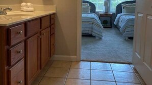 Een badkamer met een wastafel, spiegel en houten kasten. Een deur leidt naar een slaapkamer met twee bedden en een raam.