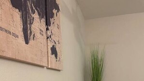 Een kamer met een donker houten vloer, een kamerkleed met dessin, een zwarte leren bank, een hoge groene plant in een metalen vaas, en twee wereldkaarten aan de muur.