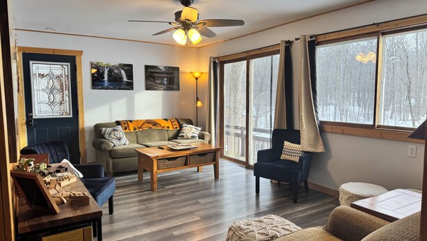 A cozy living room with a wooden floor, a ceiling fan, a sofa, a coffee table, and large windows with curtains. - Charming 2-bedroom cottage with WiFi, AC on quiet lake on trails with kayaks. (Birchwood)