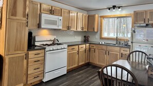 A modern kitchen with wooden cabinets, a white stove, a microwave, and a window with a view of trees. - Charming 2-bedroom cottage with WiFi, AC on quiet lake on trails with kayaks. (Birchwood)