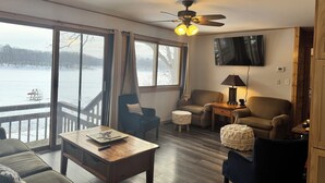 A cozy living room with a large window, a ceiling fan, a television, and a fireplace. - Charming 2-bedroom cottage with WiFi, AC on quiet lake on trails with kayaks. (Birchwood)