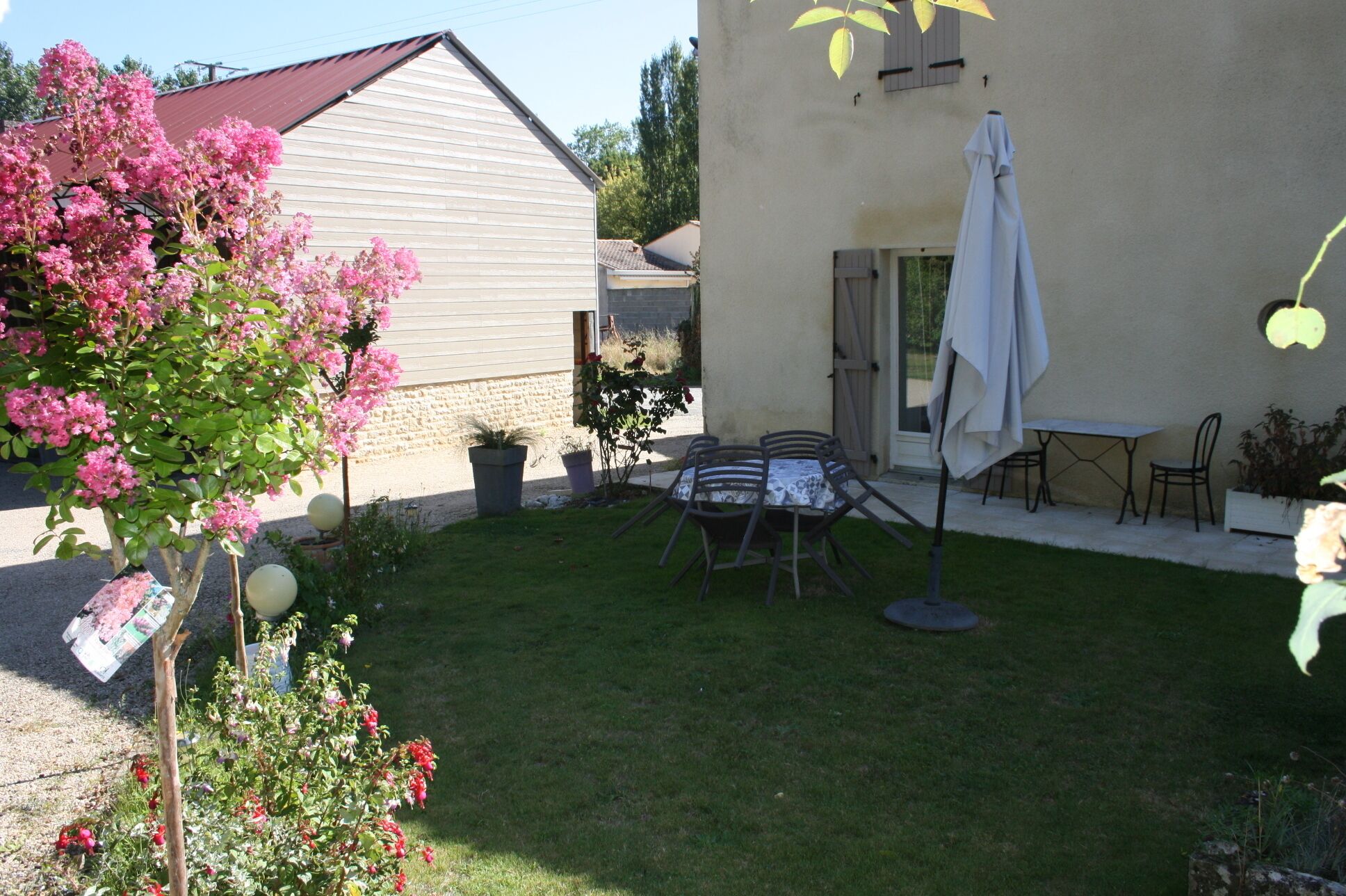 Eine Terrasse mit Tisch und Stühlen, einem weißen Sonnenschirm und einem Garten mit rosa Blumen.