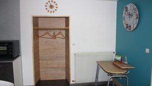 Una stanza modesta con una porta in legno, un termosifone, un tavolo con una sedia e una sveglia appesa al muro.