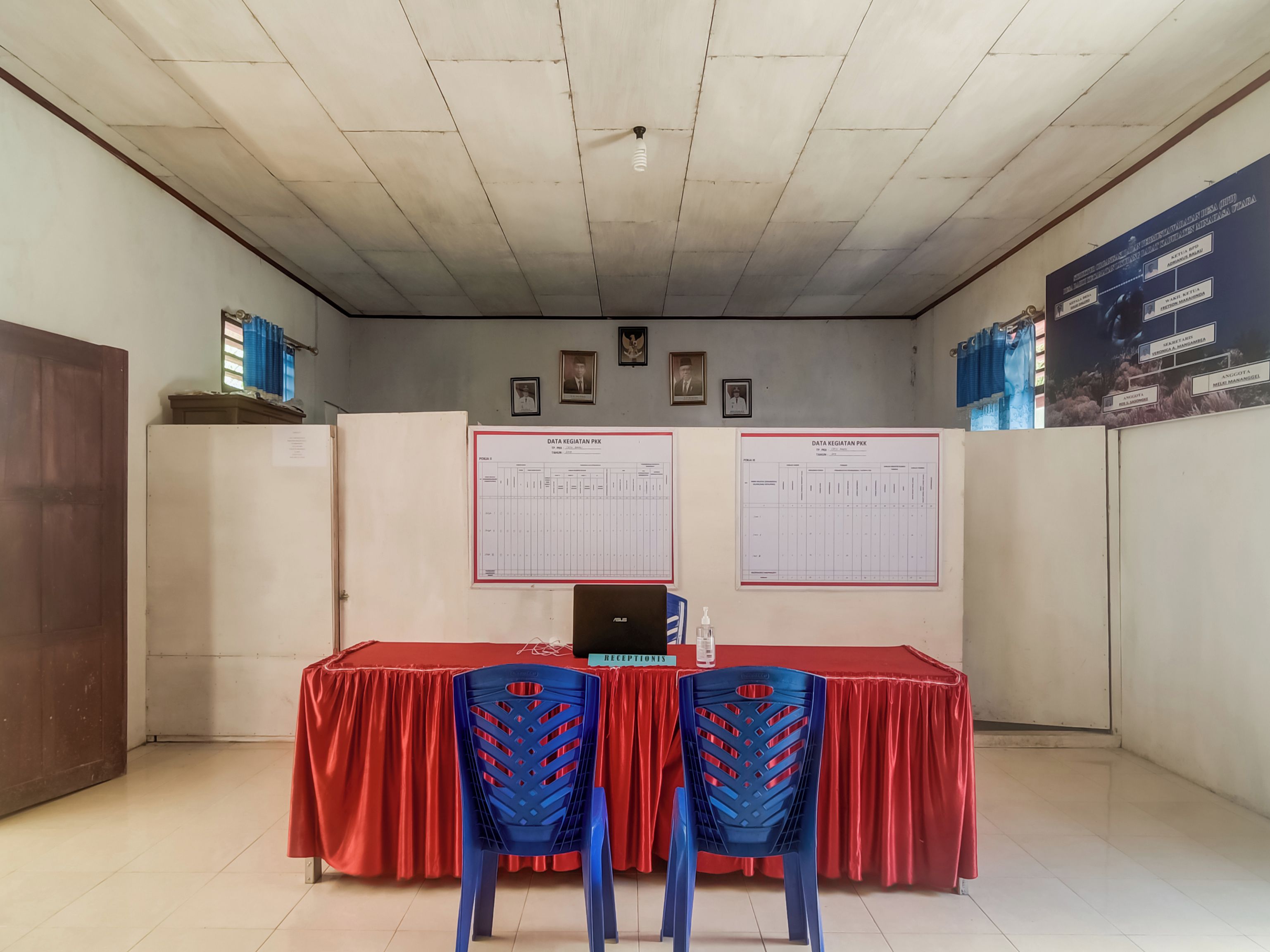 Una habitación con mostrador de recepción, dos sillas azules, una computadora portátil y dos carteles grandes en la pared.