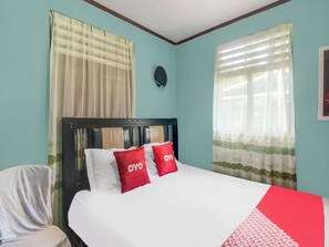 Ein Bett mit weißer Bettwäsche und roten Kissen, ein Stuhl mit weißem Bezug und ein Fenster mit transparenten Vorhängen.
