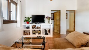 Uma sala de estar com televisão, prateleiras com objetos decorativos, uma mesa de centro de vidro e um sofá.