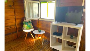 Una habitación de madera con una TV sobre un mueble blanco, una mesita con una silla y una ventana con cortinas.