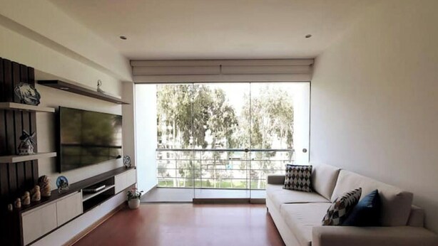 Uma sala de estar moderna com televisão de tela plana, um sofá e uma varanda com vista para árvores.