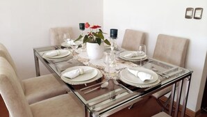 Mesa de jantar posta para seis com tampo de vidro, pratos brancos e guardanapos. Há uma planta em vaso e velas sobre a mesa.