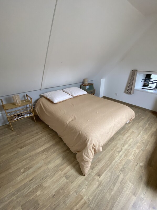 Quarto com cama, piso de madeira, uma pequena mesa de cabeceira, prateleira na parede e janela com persianas.