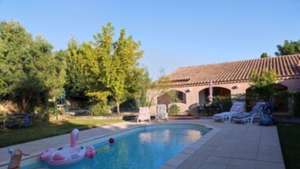 Una piscina con juguetes inflables, un área de patio con sillones de descanso y un edificio con techo de tejas.