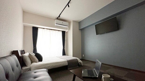 A hotel room with a sofa, a bed, a coffee table with a laptop, a TV mounted on the wall, and an air conditioning unit. - Nagomi by b hotel | Comfortable Studio Stay N22 (Hiroshima)