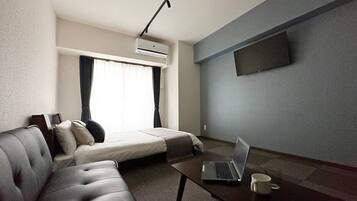 A hotel room with a sofa, a bed, a coffee table with a laptop, a TV mounted on the wall, and an air conditioning unit.