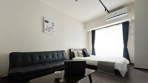 A hotel room with a black sofa, a bed with white bedding, a small table with a glass, and a wall-mounted air conditioner. - Nagomi by b hotel | Comfortable Studio Stay N22 (Hiroshima)