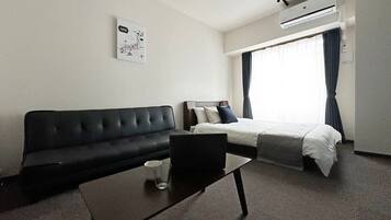 A hotel room with a black sofa, a bed with white bedding, a small table with a glass, and a wall-mounted air conditioner.