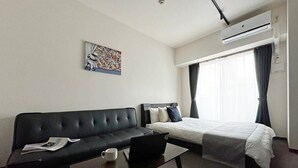 A hotel room with a black sofa, a bed with white bedding, a small table with a laptop and a cup, and an air conditioning unit on the wall.