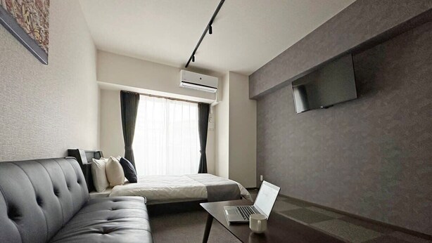 A modern hotel room with a sofa, a bed, a flat-screen TV mounted on the wall, and a laptop on a small table.