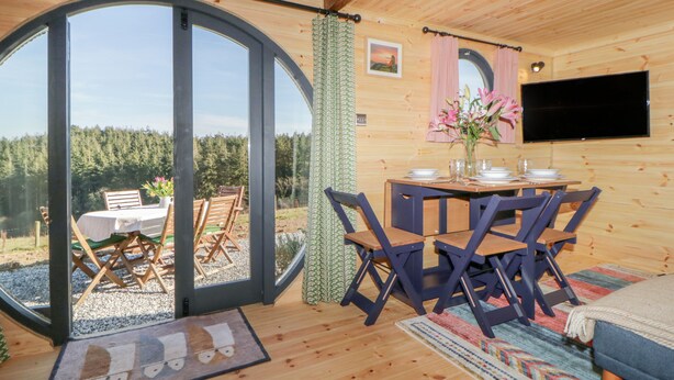 A cozy cabin interior with a dining area, a TV mounted on the wall, and a large sliding glass door leading to a scenic outdoor view.