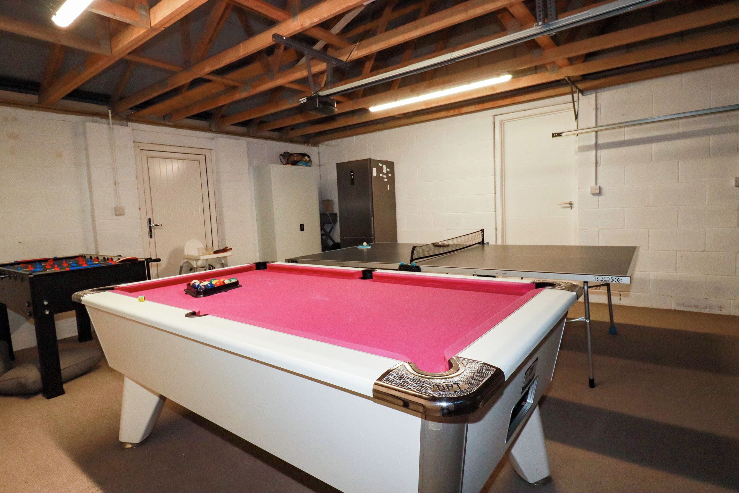 Una mesa de billar con superficie de fieltro rosa, una mesa de fútbol de mesa y una mesa de ping pong en una sala con paredes blancas y vigas de madera en el techo.