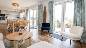 A modern living room with a dining area, a large window with curtains, and a surfboard on the wall. - Lavender House (Newquay)