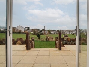 A view through a glass door leading to a courtyard with a grassy area, wooden posts, and a distant village. - Lavender House (Newquay)