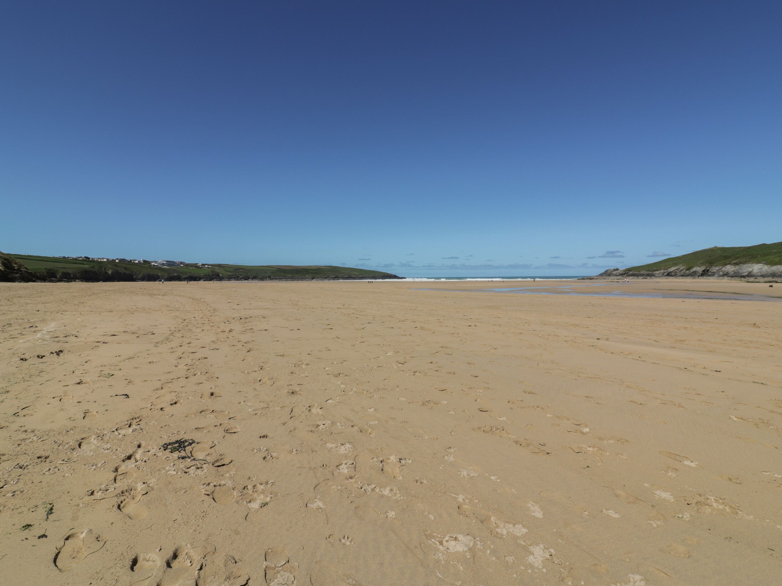 A wide sandy beach with footprints, grassy hills in the background, and a clear blue sky.