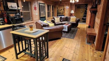 A cozy cabin interior with a wooden floor, a dining area with a table and bar stools, a leather sofa, and a kitchen area with a microwave.