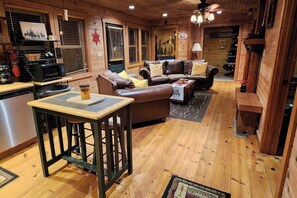 Acogedor interior de cabaña con suelo de madera, zona de comedor con mesa y taburetes de bar, sofá de cuero y área de cocina con microondas.