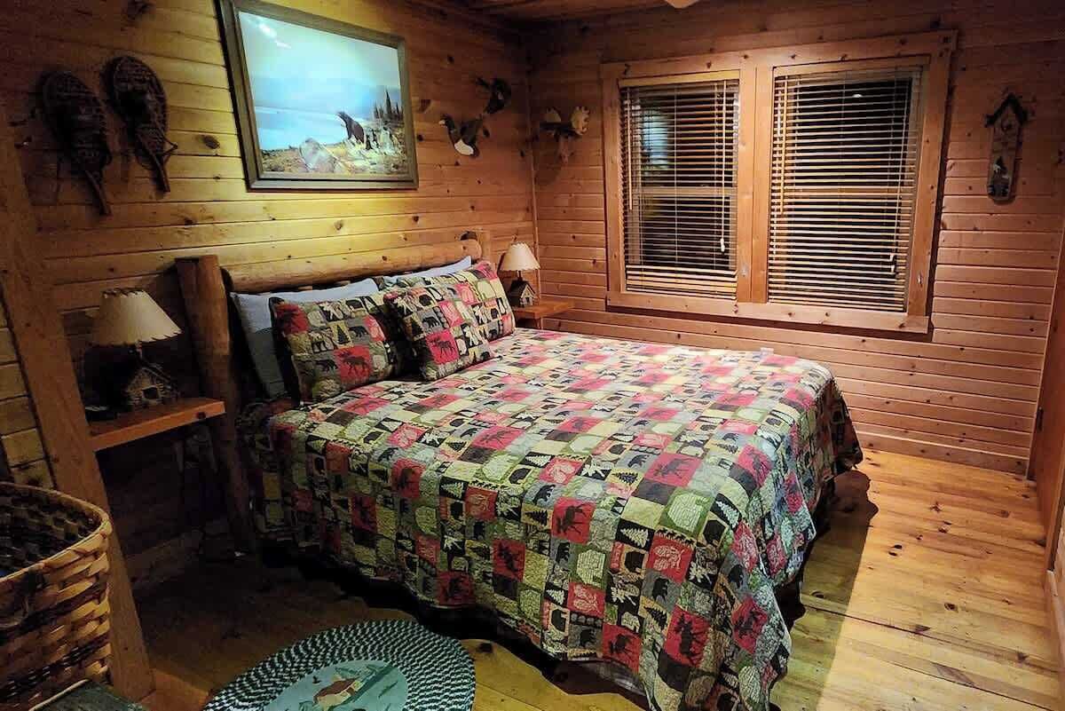 A cozy wooden cabin bedroom with a bed, a window with blinds, and wall decorations including animal heads.