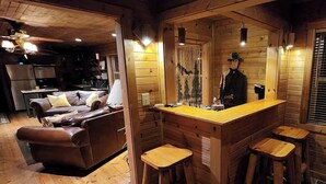 Ein gemütliches Blockhaus-Interieur mit einem Wohnbereich, der eine braune Ledersofa, eine Holzbar mit Hockern und eine Küche im Hintergrund zeigt.